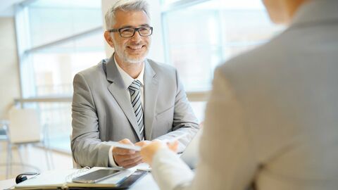 Beratung Älterer Mann mit einem grauen Anzug sitzt einer Person gegenüber und gibt dieser ausgedruckte Unterlagen.